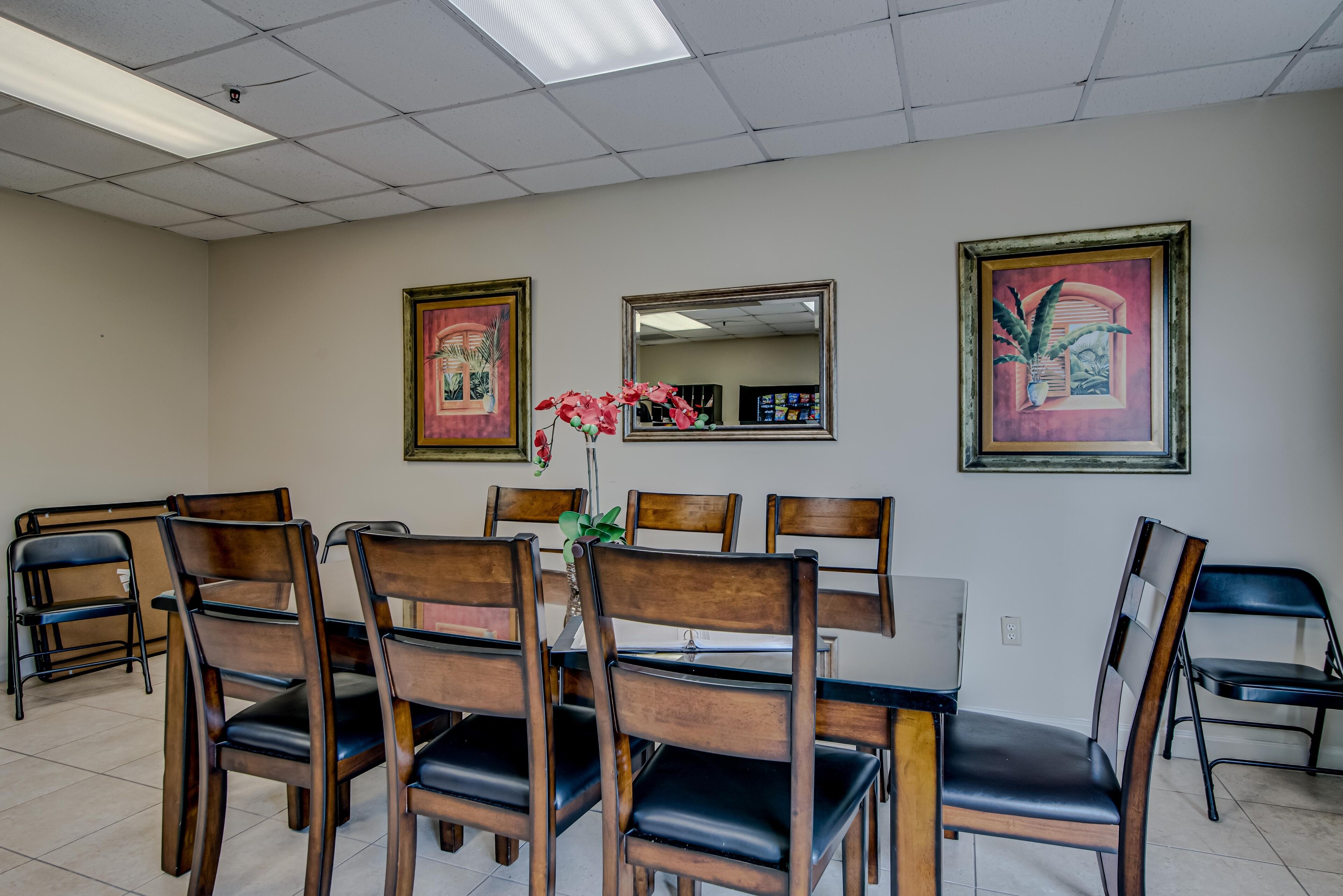 A conference room with a rectangular table surrounded by wooden chairs with black cushions. There are three framed artworks on the walls and a potted plant with pink flowers on the table.