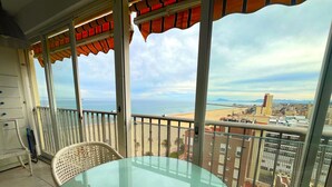 Un balcón con mesa de vidrio y silla de mimbre, con vistas a una playa y al paisaje urbano.