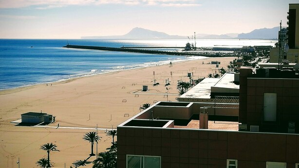 Una playa con un muelle que se adentra en el océano, cielo despejado y edificios al frente.