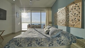 Un dormitorio con una cama grande, un ventilador de techo, vista del cielo a través de una ventana y un panel decorativo en la pared.