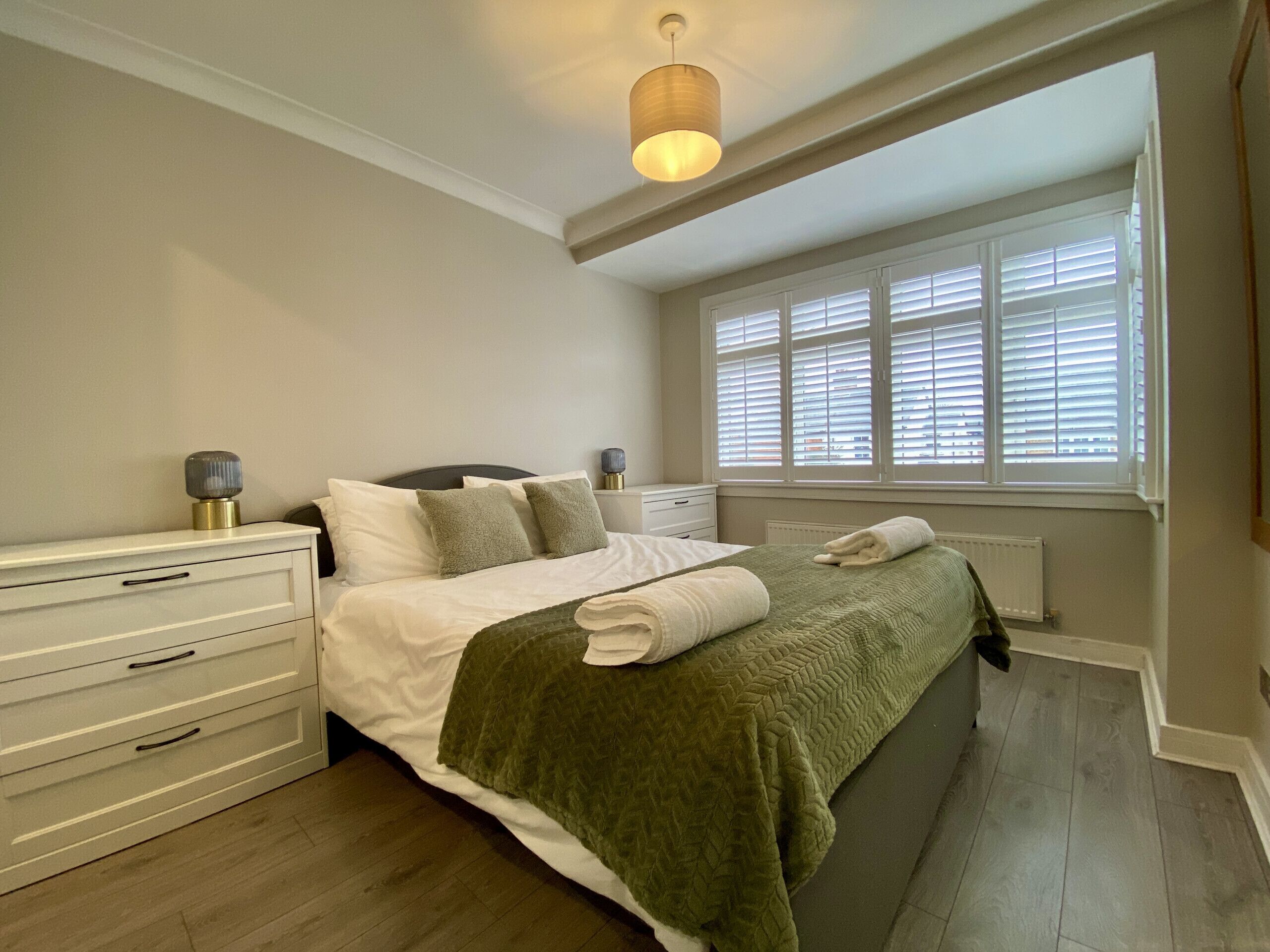 Une chambre à coucher comprenant un lit, des tables de chevet, une fenêtre avec des stores et un luminaire au plafond.