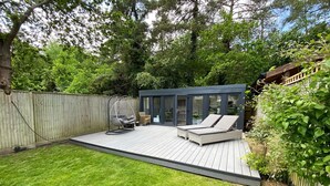 A backyard with a wooden deck, two lounge chairs, a hanging chair, and a glass-enclosed patio area.