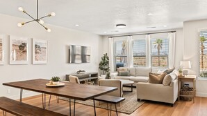 A modern living room with a wooden dining table, a sofa, and a TV mounted on the wall. - Sandstone Shores: Desert Color Lagoon + Beaches (St. George)