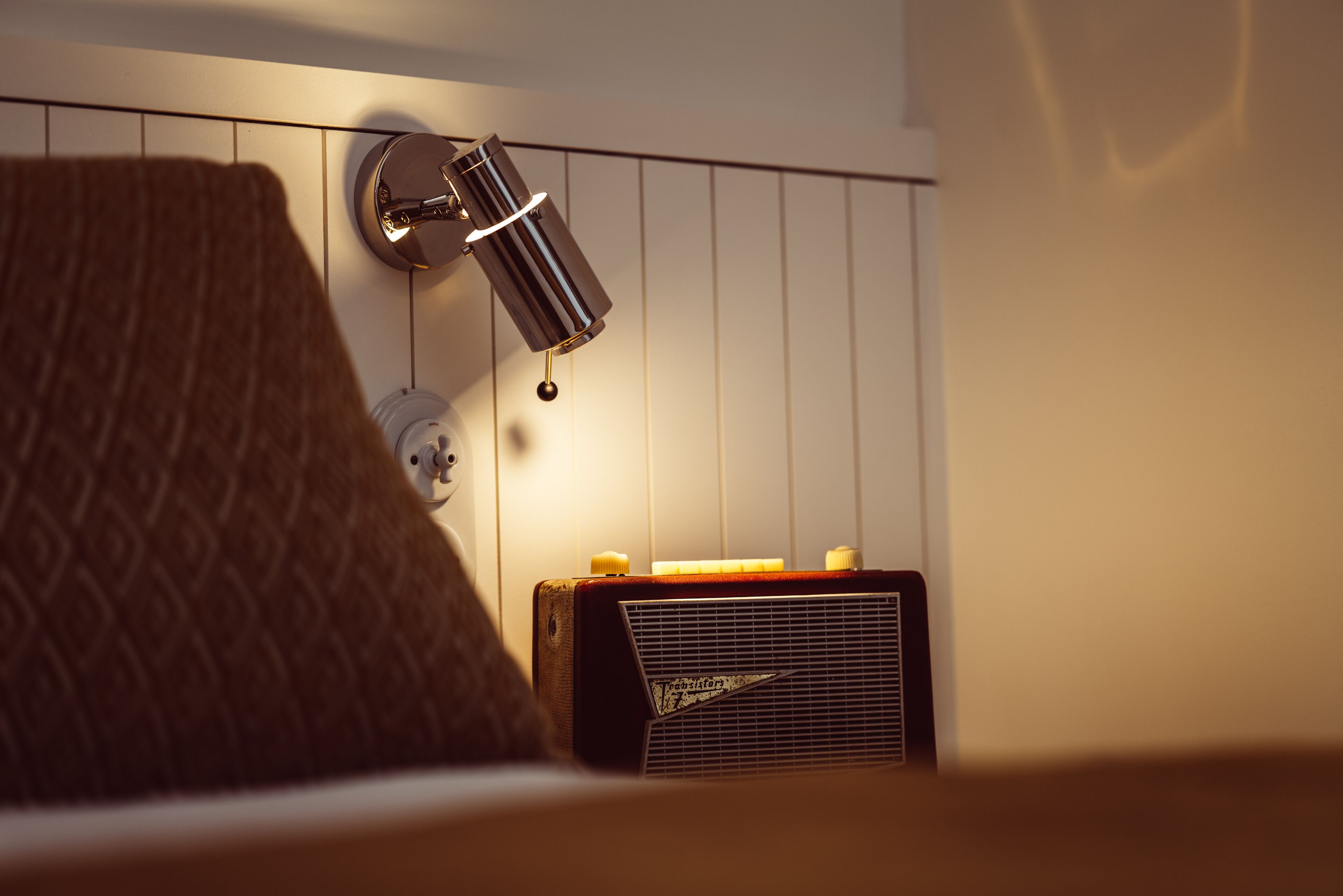 Una habitación con un cabecero marrón estampado, una radio antigua y una lámpara fijada a la pared.
