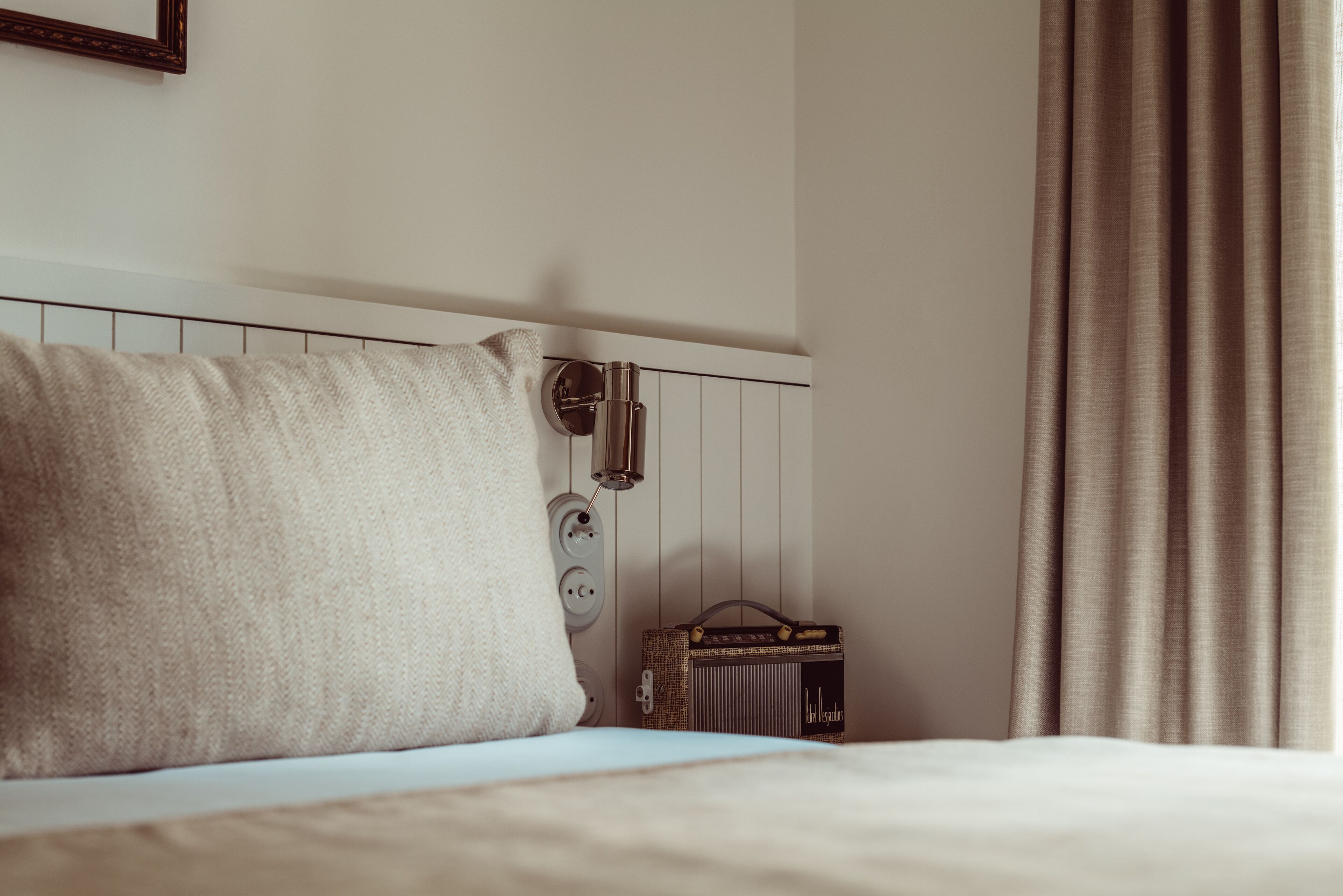A neatly made bed with a pillow, a vintage radio, and a power outlet on the wall.