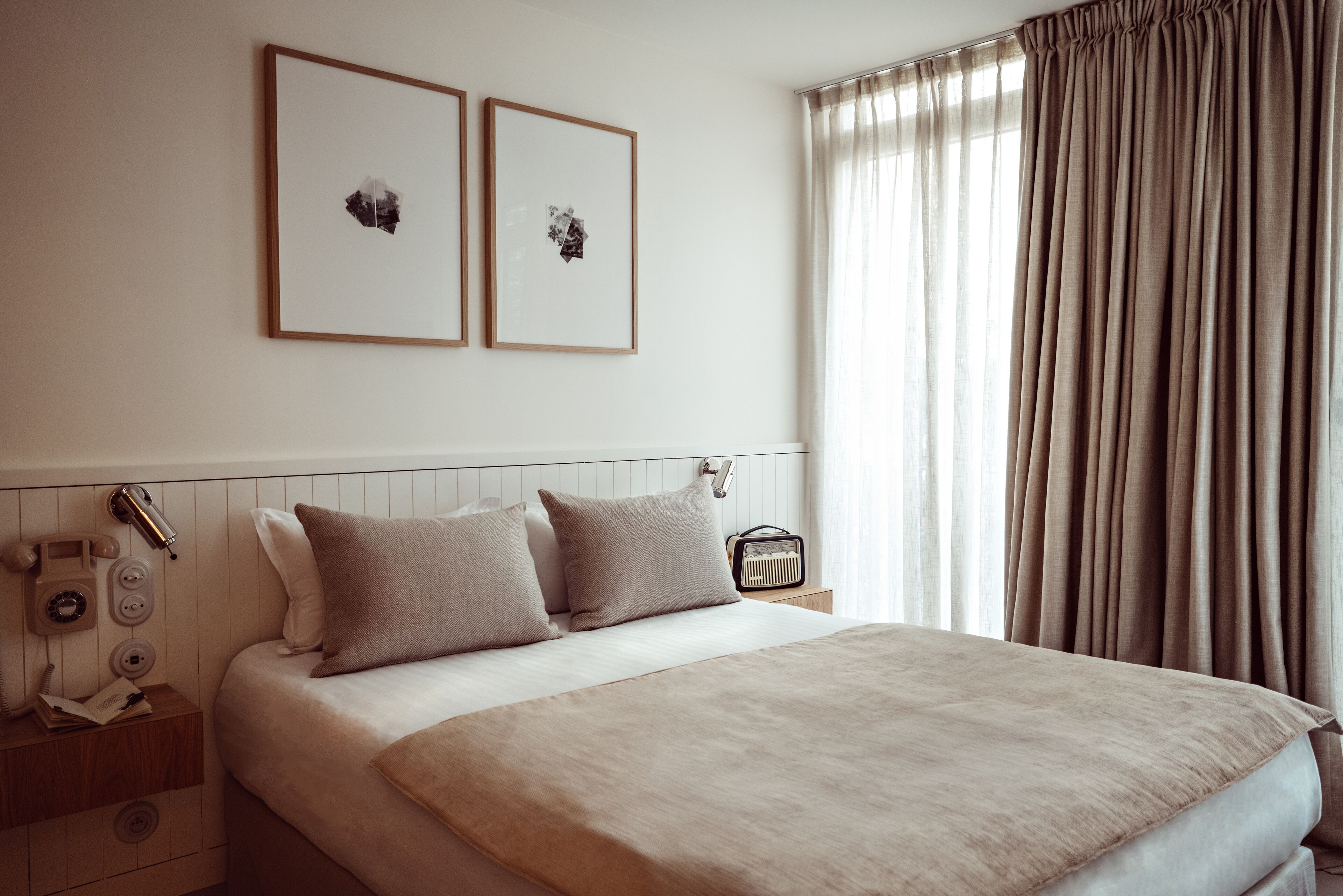 Ein ordentlich eingerichtetes Schlafzimmer mit einem Bett, zwei Kissen, einem Nachttisch mit Lampe, einem Radio und gerahmten Bildern an der Wand.