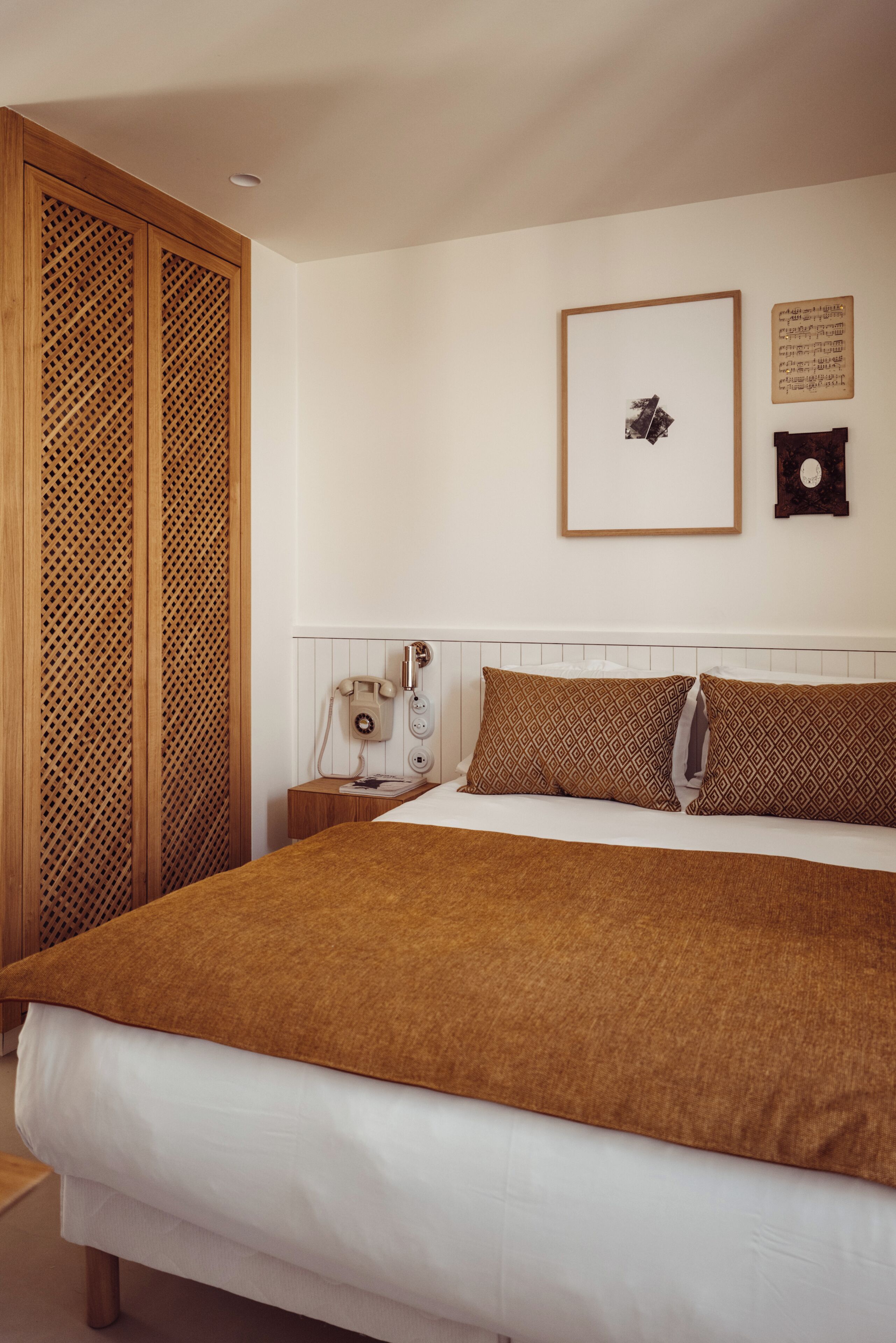 A bedroom with a bed, wooden wardrobe, bedside table, and a wall-mounted telephone.