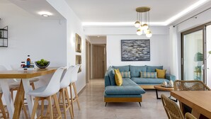 A modern living room with a sofa, dining table, and chairs. There is a painting on the wall and a ceiling light fixture.