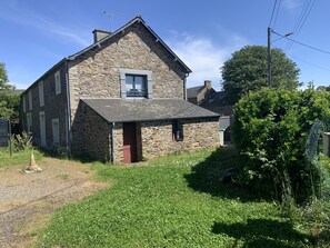 Situated in a rural setting, this traditional house features a small porch and a well-maintained lawn with shrubs and trees.