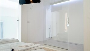 Un baño moderno con ducha envuelta en vidrio, una cama con toallas ordenadas y un panel de control a la vista.