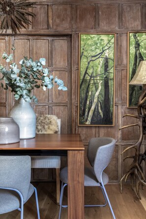 A wooden dining area with a table, chairs, a vase with foliage, a painting of a forest, and a decorative lamp. - Cockspur Street by onefinestay (London)
