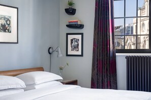A hotel room with a bed, a wall-mounted lamp, framed artwork, a small plant, and a view of the city through a window.