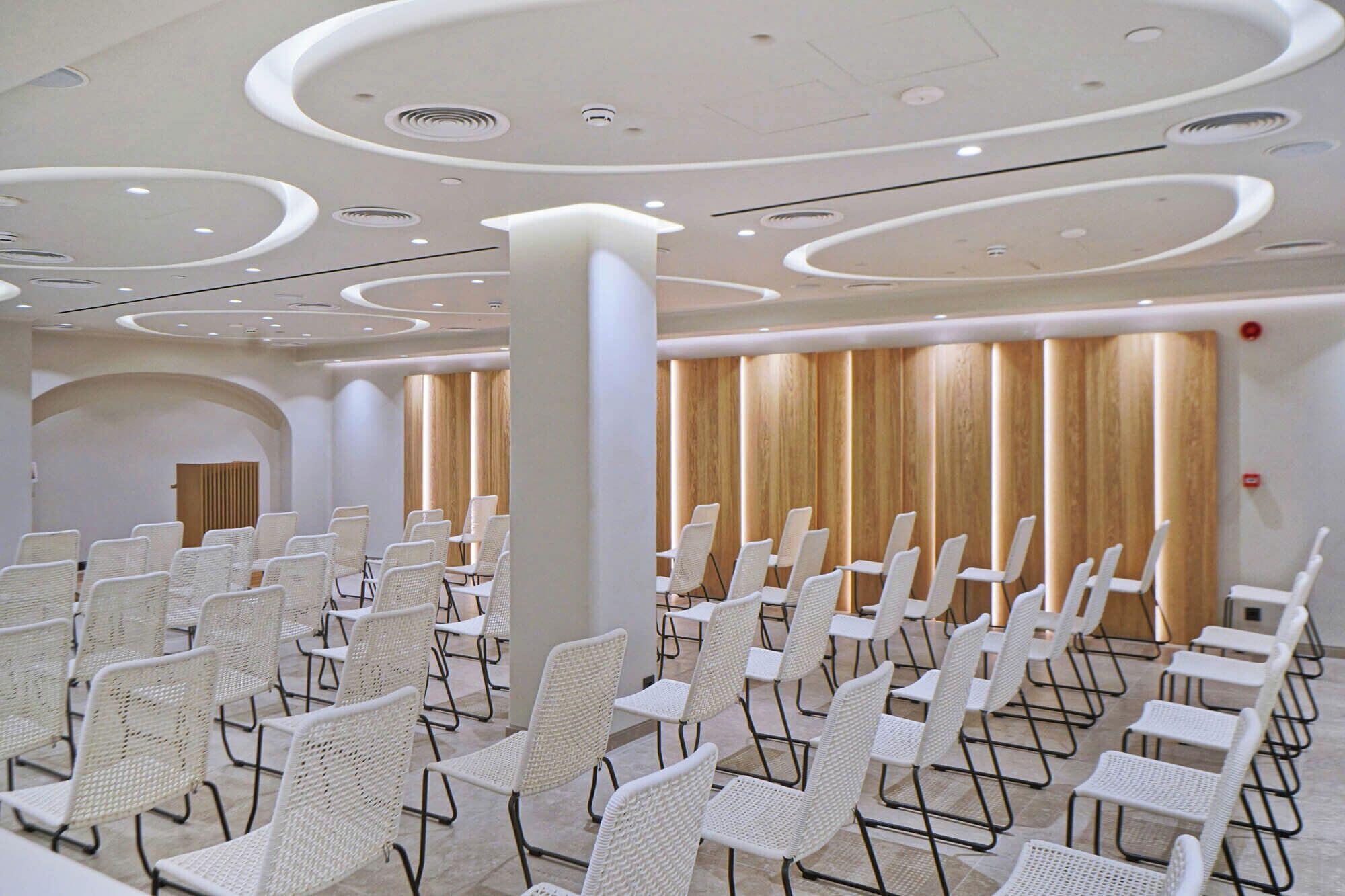 A conference room with white chairs arranged in rows, a wooden panel wall, and a modern ceiling design.
