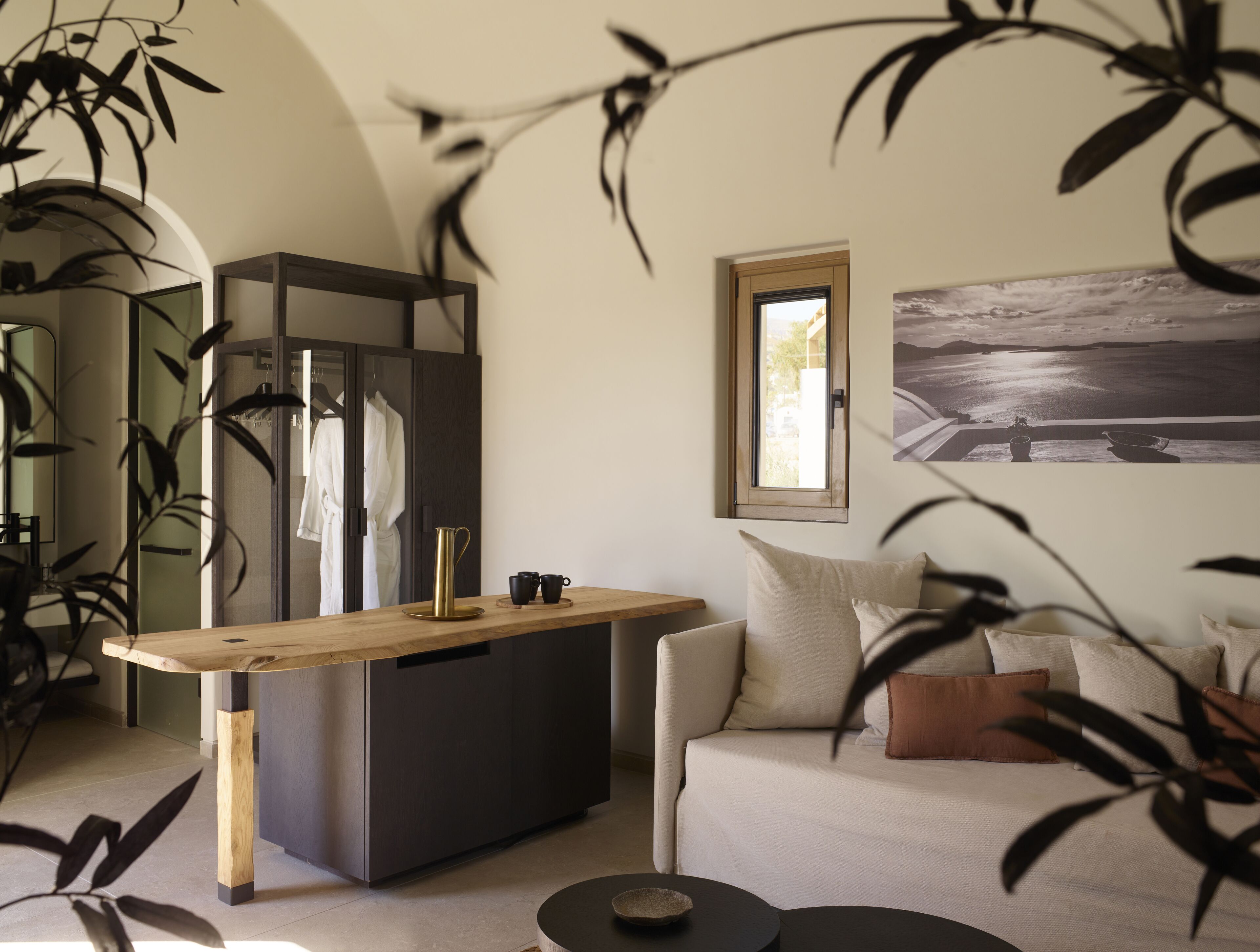 A modern living room with a wooden coffee table, a sofa with cushions, a large mirror, and a view of the sea through a window.