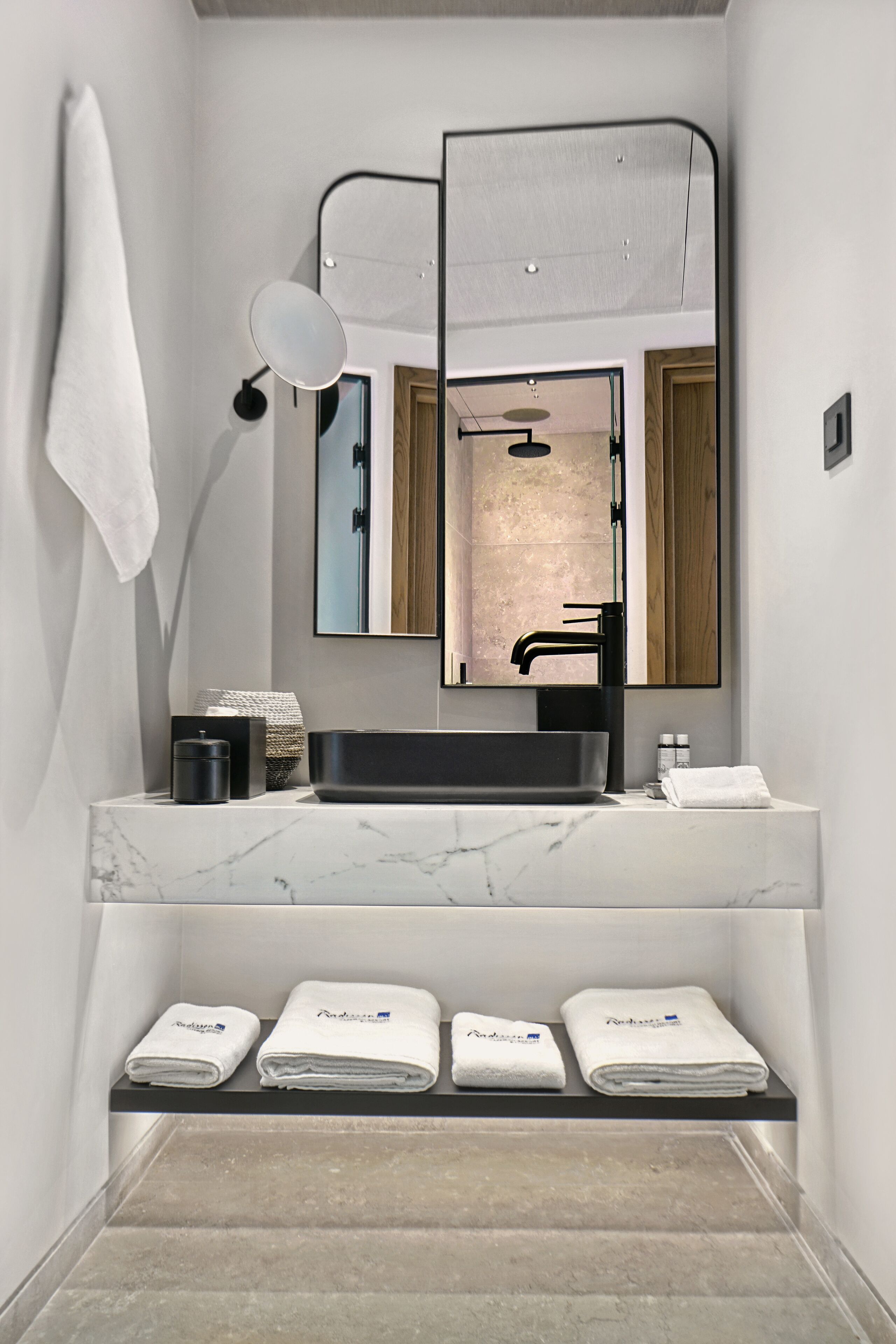 A modern bathroom with a marble countertop, a black sink, and a large mirror. There are three white towels on a shelf below the sink.