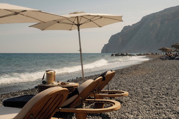 Une plage avec des chaises longues en bois et des parasols, un rivage rocheux et une falaise en arrière-plan.