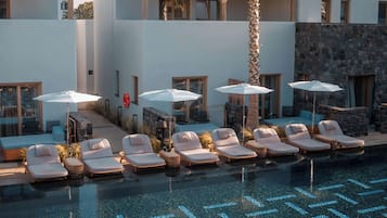 A poolside area with lounge chairs and umbrellas, a stone wall, and a view of mountains.