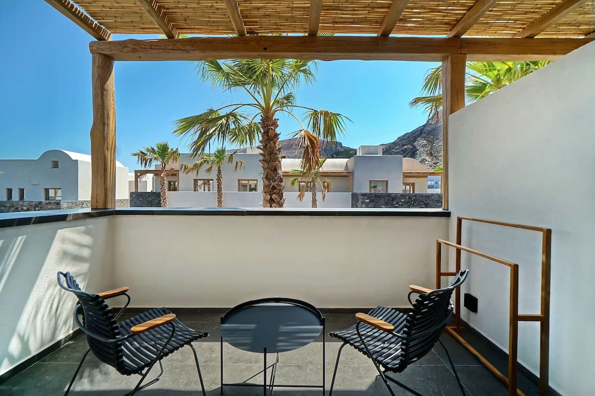 A balcony with two chairs and a table, overlooking a coastal area with palm trees and buildings.
