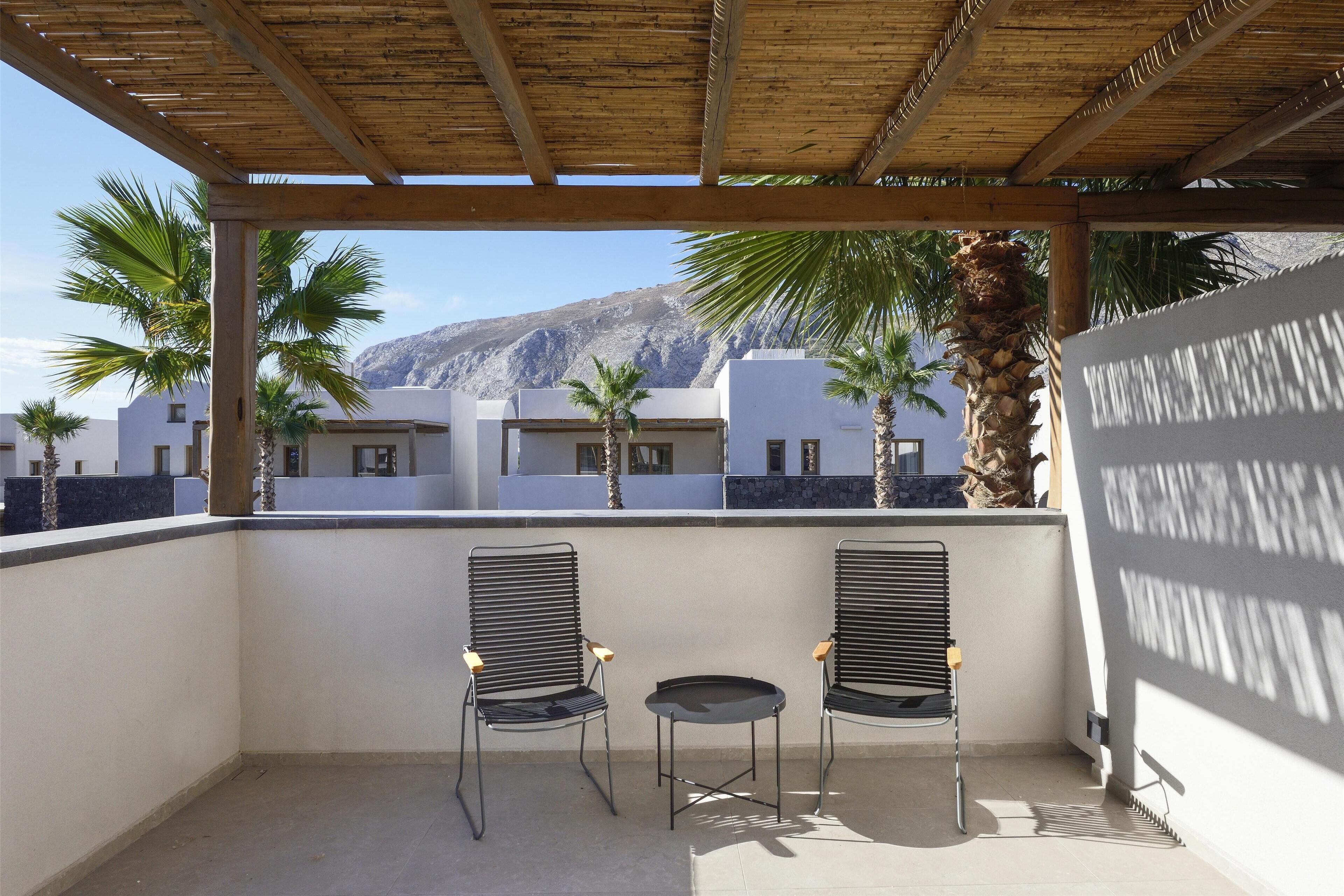 A balcony with two chairs and a small table, overlooking a mountain and palm trees.
