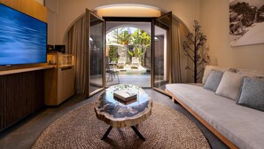 A modern living room with a large flat-screen TV, a round coffee table, and a sofa. There is a glass door leading to an outdoor area with a view of greenery and a stone wall.