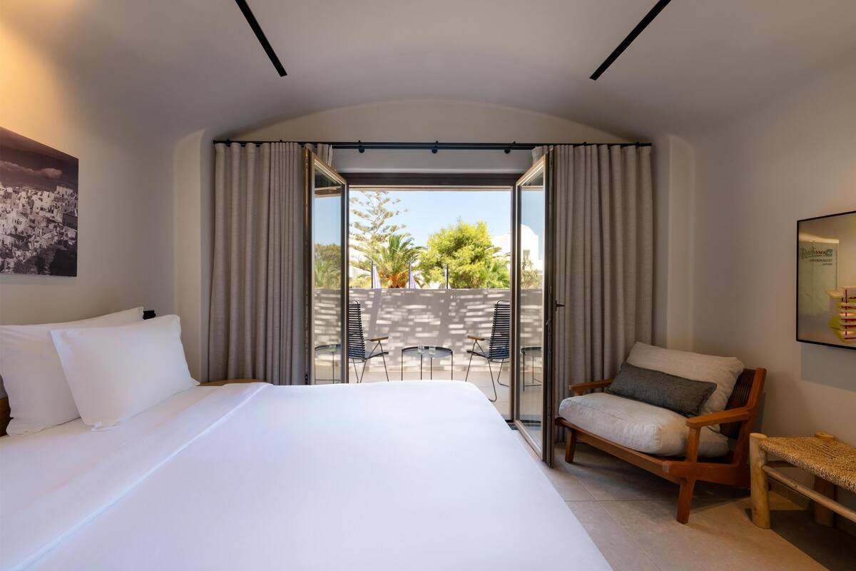 A hotel room with a large bed, a wooden chair, a small table, and a balcony with curtains.