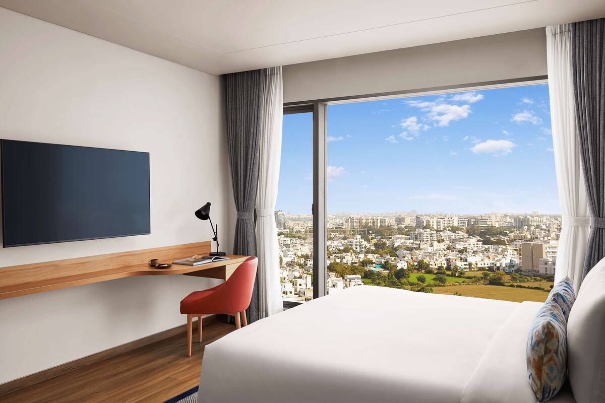 Habitación de hotel moderna con una cama grande, un escritorio con silla, televisor de pantalla plana y vista a la ciudad a través de amplios ventanales.