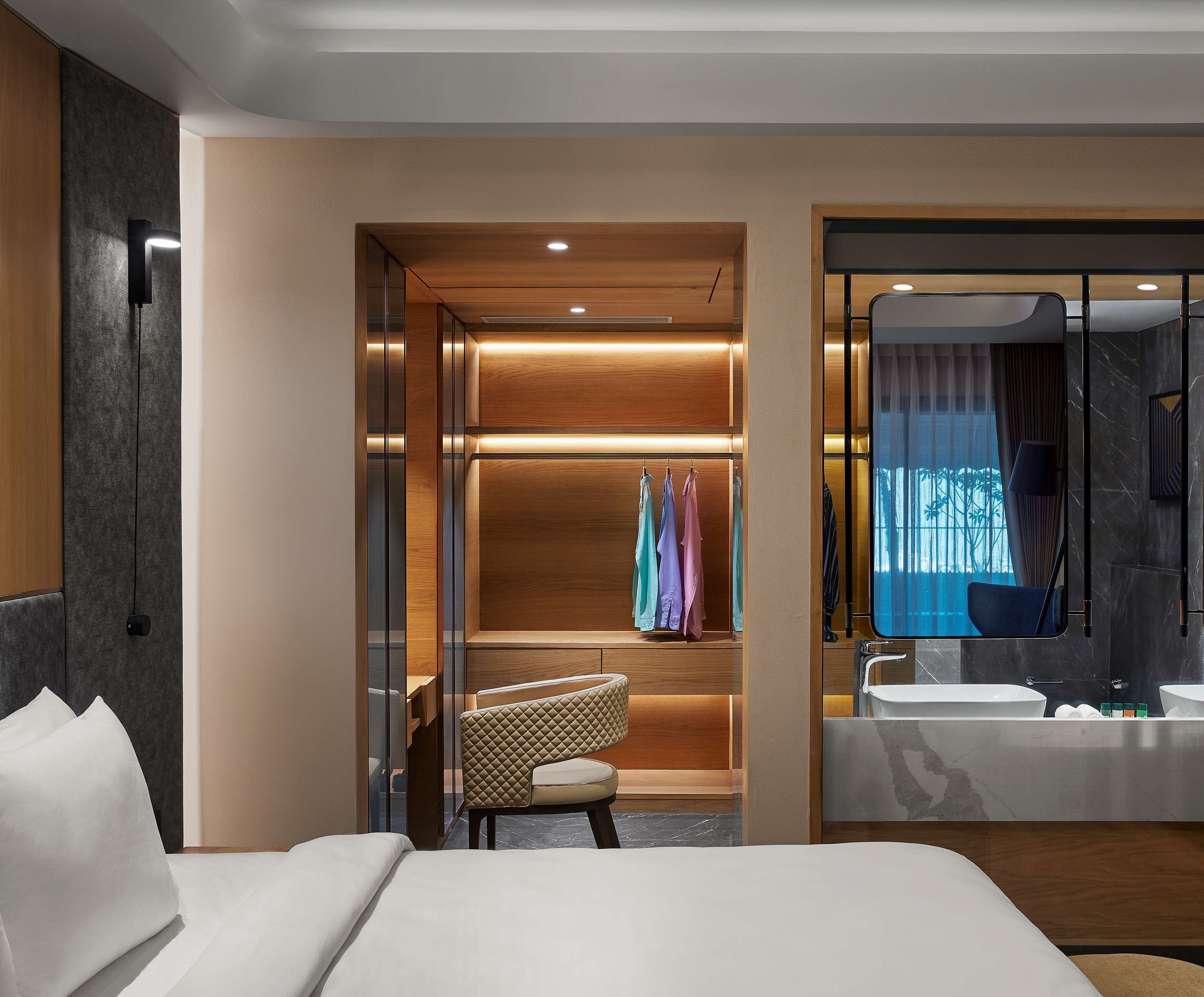 A modern hotel room with a bed, a seating area, a walk-in closet, and a bathroom with a large mirror and sink.