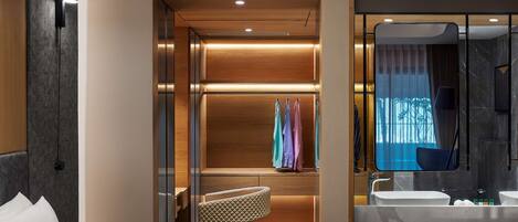 A modern hotel room with a bed, a seating area, a walk-in closet, and a bathroom with a large mirror and sink.