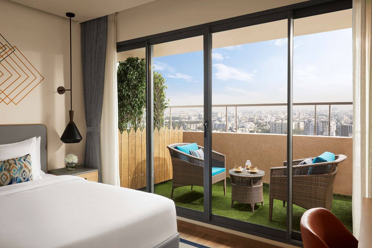 Una habitación de hotel con una cama grande, un balcón con mobiliario de mimbre y vistas a la ciudad.