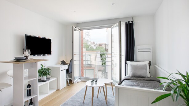 A modern, well-lit room with a bed, a desk, a TV, and a view of the city through large windows.
