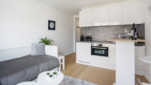 A modern, compact living space with a kitchenette, a bed with grey bedding, a small white table, and a potted plant.
