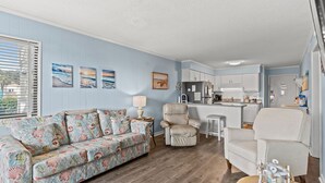 A living room with a sofa, chairs, and a small table. There is a kitchen area with cabinets and a countertop. - Ocean Forest Villas 114A - Charming 2 Bed/2 Bath Condo in Myrtle Beach, Outdoor pool, Kiddie Pool (Myrtle Beach)