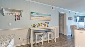 A kitchen with a white countertop, two stools, a shelf with a 'BEACH' sign, and a painting of a beach. - Ocean Forest Villas 114A - Charming 2 Bed/2 Bath Condo in Myrtle Beach, Outdoor pool, Kiddie Pool (Myrtle Beach)