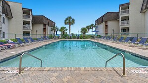 A swimming pool with lounge chairs and palm trees, surrounded by apartment buildings. - Ocean Forest Villas 114A - Charming 2 Bed/2 Bath Condo in Myrtle Beach, Outdoor pool, Kiddie Pool (Myrtle Beach)