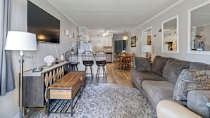 Un salon moderne avec un canapé, une table à manger en bois et des chaises. Un téléviseur est fixé au mur, ainsi qu’une lampe et un tapis à motifs.