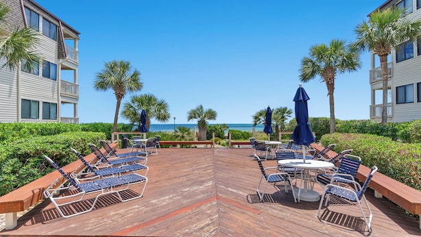 Une terrasse en bois, dotée de chaises longues et de parasols, qui offre une vue sur une plage bordée de palmiers et un ciel d’un bleu limpide.