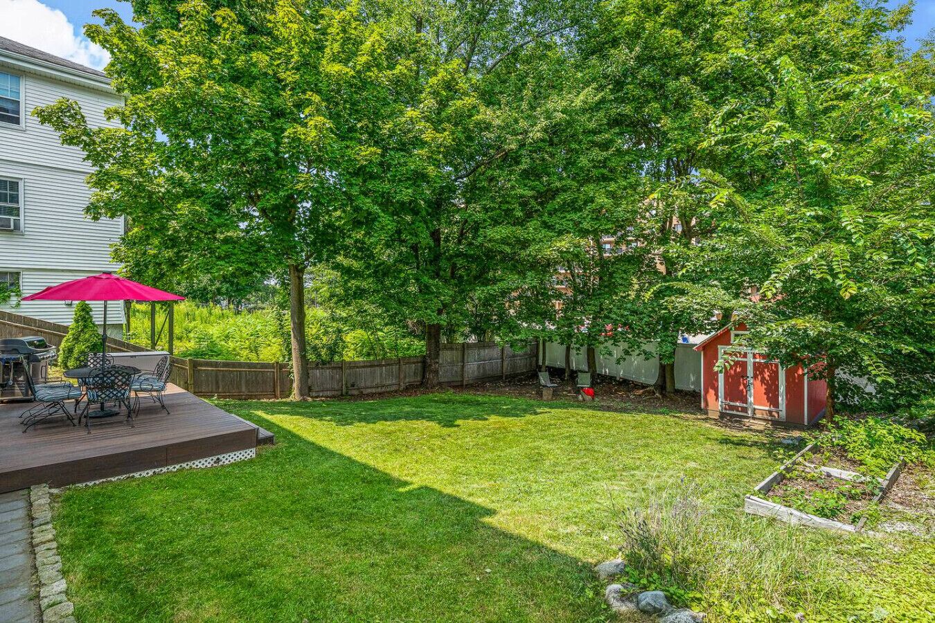 A backyard with a wooden deck, a red umbrella, a grill, and a shed.