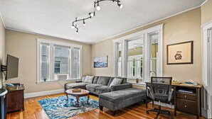 A living room with a grey sectional sofa, a wooden coffee table, a desk with a chair, and a blue patterned rug. - Next to Boston & Beach, King Beds, Free Parking+EV (Quincy)