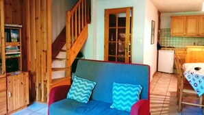 A cozy living room with a sofa, a wooden staircase, a kitchen area, and a small table with a tablecloth. - Clos des Loirs, 6. 55m2 duplex apartment, not overlooked (Samoëns)