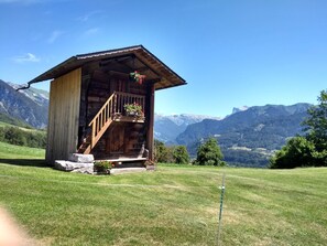 Una cabaña de madera con balcón, rodeada por un campo cubierto de hierba y montañas al fondo.