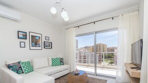 A modern living room with a white sofa, a round wicker coffee table, and a large window offering a city view. - Emerald Apartment, AC, Swimming Pool !new! (Portimão)