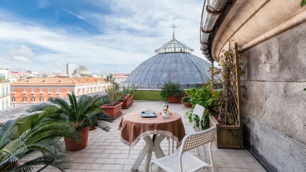 A terrace with a round table and chairs, potted plants, and a view of a dome-shaped building in the background.