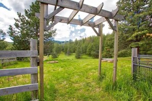 Una estructura de pérgola de madera en un entorno natural, con un campo cubierto de césped y árboles al fondo.