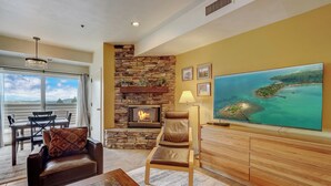 A living room with a stone fireplace, a large TV, and a dining area with a view of the ocean. - Lodge at Mtn Village B148 | Ski-In/Out | Valley View | Washer/Dryer | Pool | Hot Tub (Park City)
