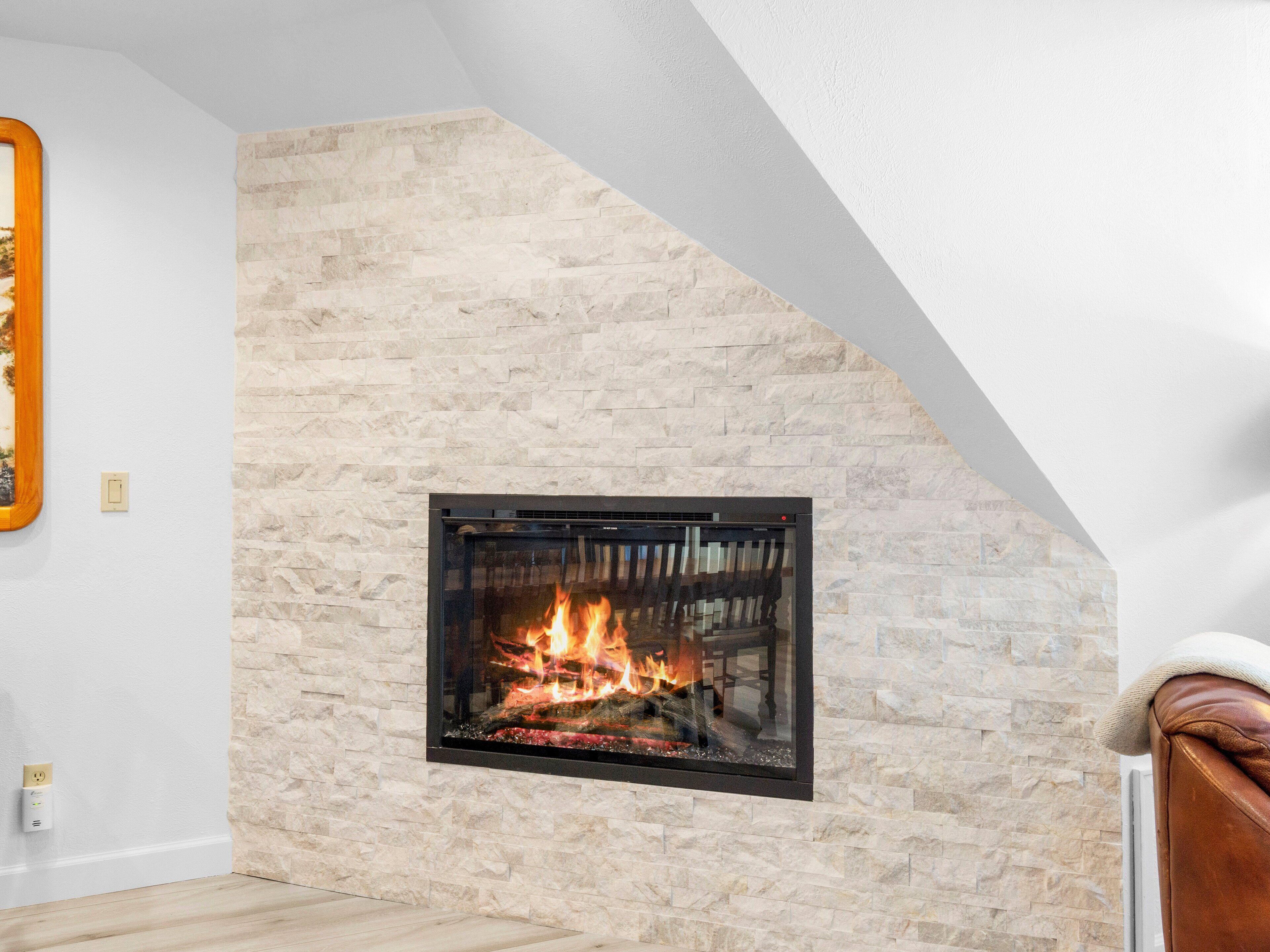 Une cheminée en pierre avec un feu de bois, une chaise en bois et un cadre photo accroché au mur.