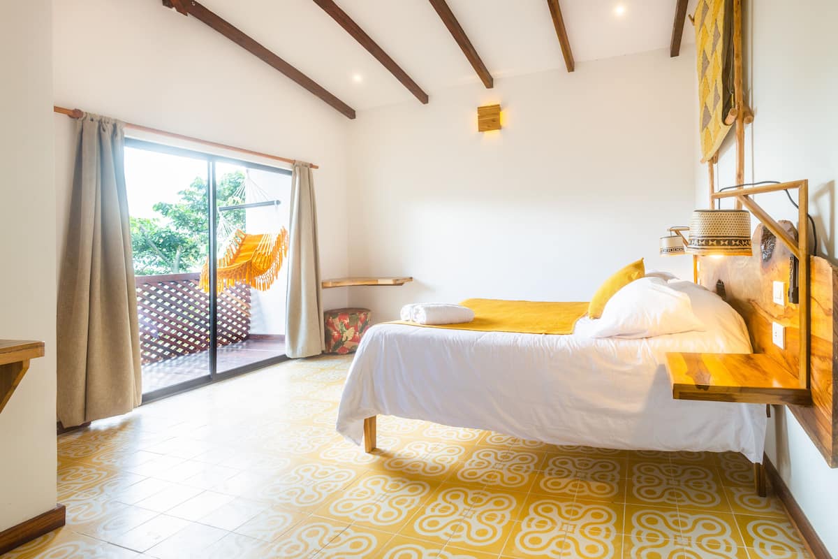 A bedroom with a bed, a wooden nightstand, a window with curtains, and a view of a balcony.
