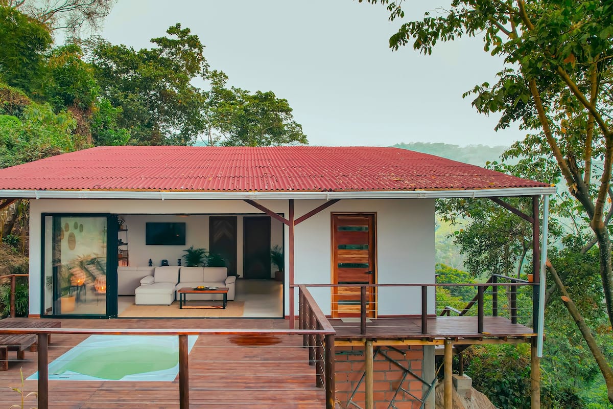 A modern house with a red roof, a glass door entrance, a living room with a sofa and coffee table, and a wooden deck with lounge chairs.