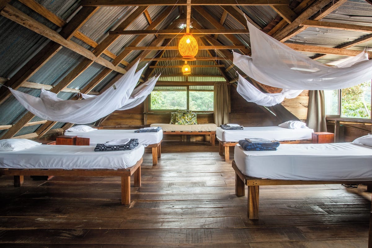 A rustic wooden room with four beds arranged in a semi-circle, a large window, and a hanging light fixture.