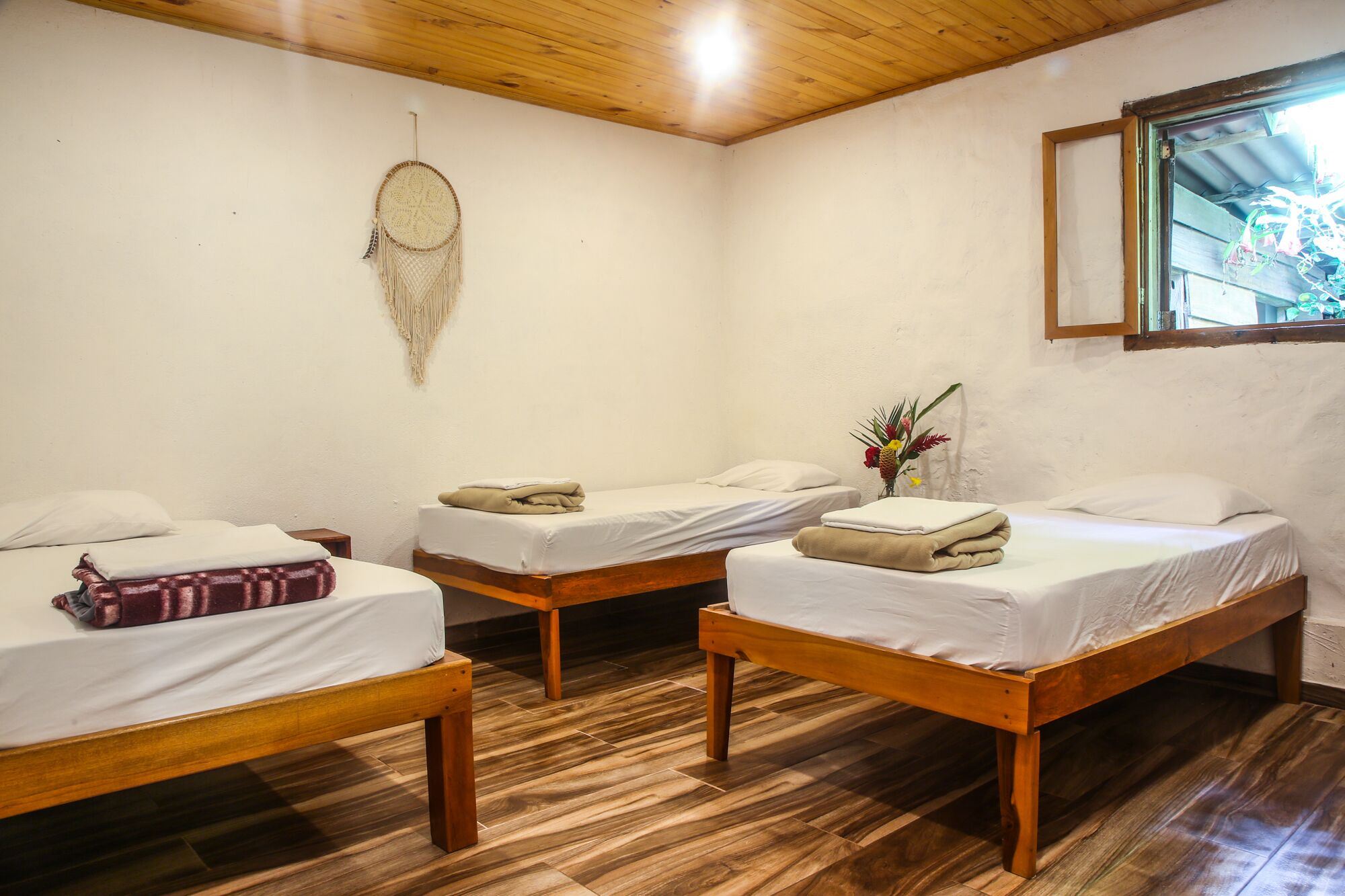 A room with three wooden beds, white bedding, a wooden floor, a dreamcatcher on the wall, and a window with a view.
