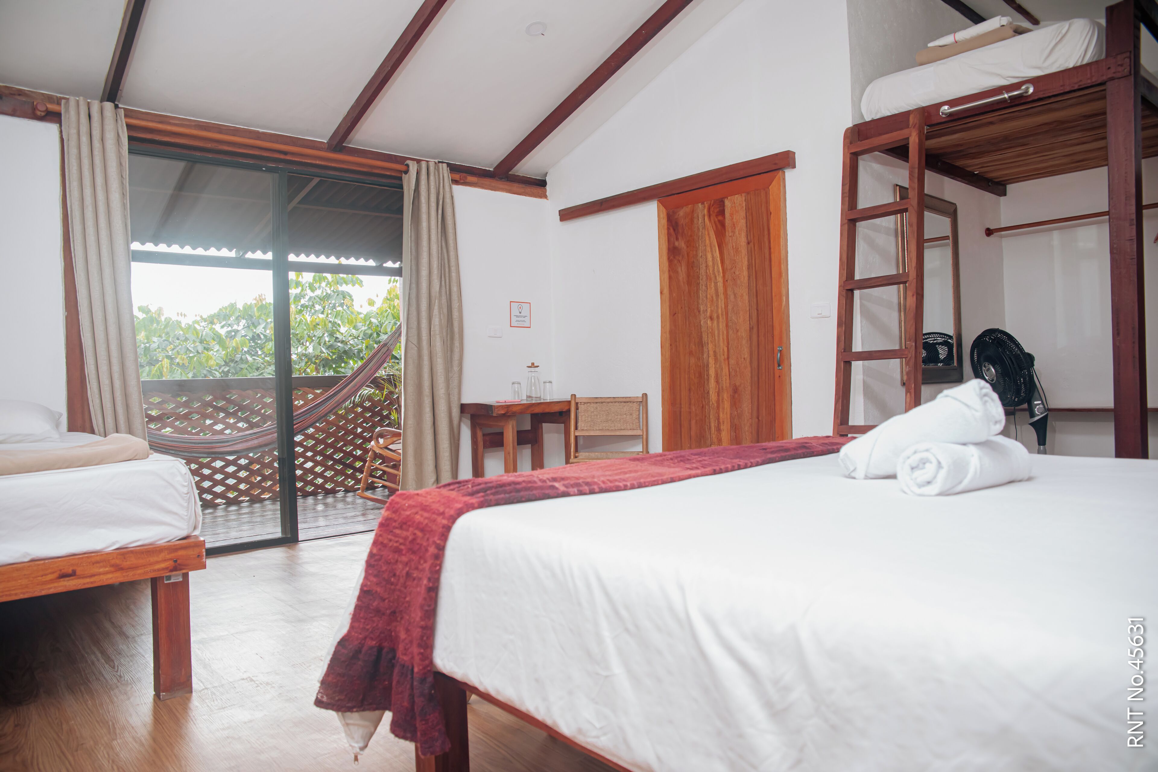 A hotel room with a bed, a wooden bench, a small table, a chair, a hammock, and a sliding glass door to the outside.